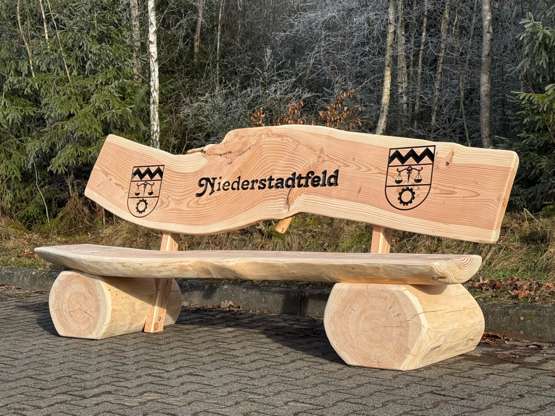 Baumstammbank mit geschwungener Baumkante, dem Schriftzug Niederstadtfeld und zwei schwarz ausgearbeiteten Gemeindewappen am Waldrand.