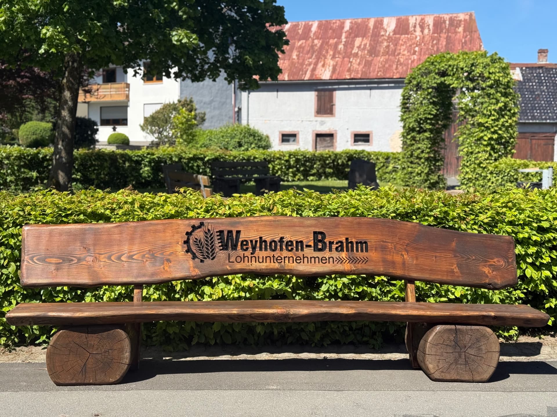Handgefertigte massive Holzbank mit detailreicher CNC-Gravur des Firmenlogos von Weyhofen-Brahm Lohnunternehmen, individuell gefertigt als repräsentative Firmenbank und langlebiger Werbeträger für den Außenbereich.