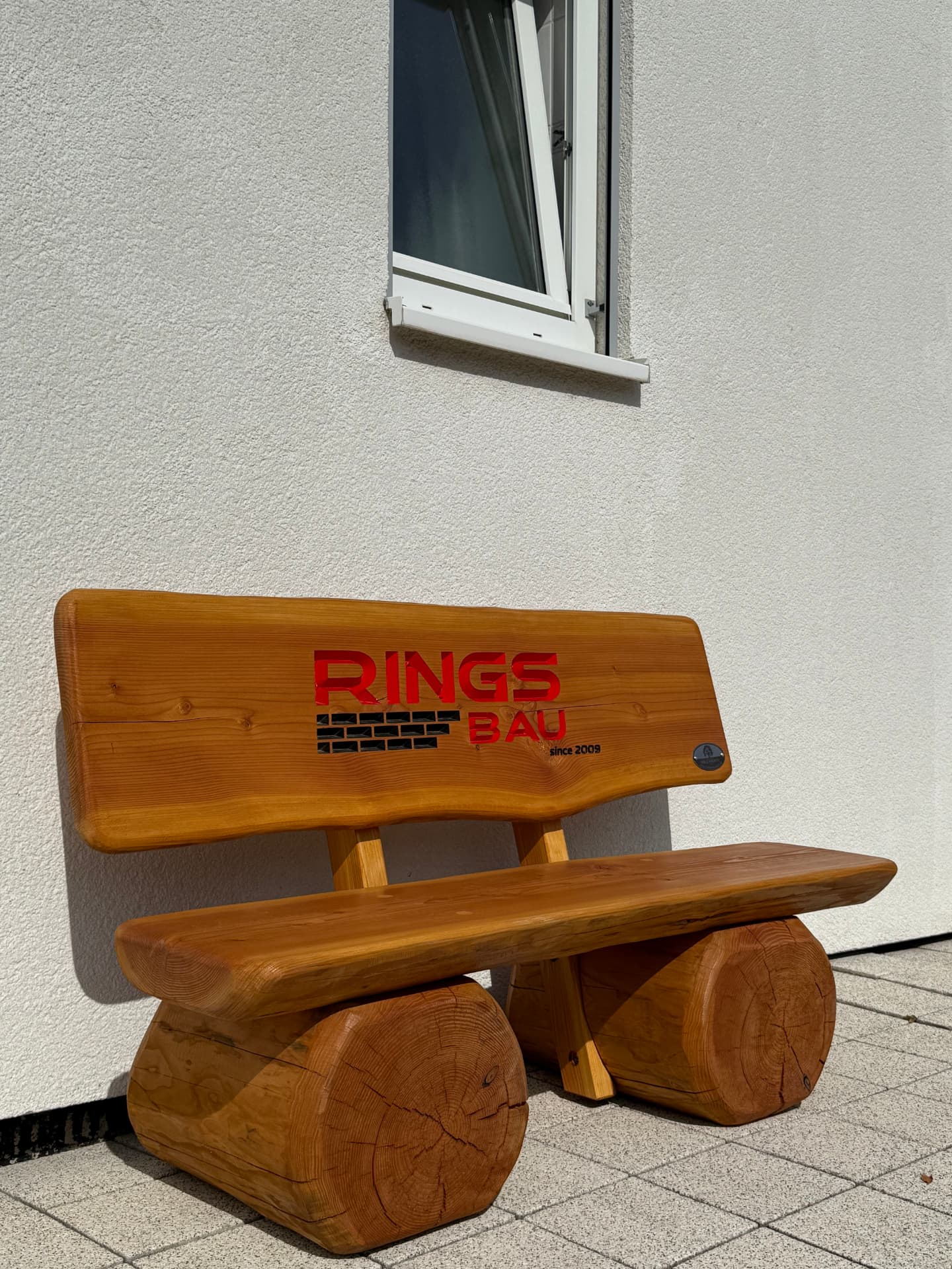Handgefertigte massive Holzbank mit farbig ausgearbeiteter CNC-Gravur des Firmenlogos von Rings Bau, individuell als repräsentative Firmenbank für den Unternehmensstandort gefertigt.