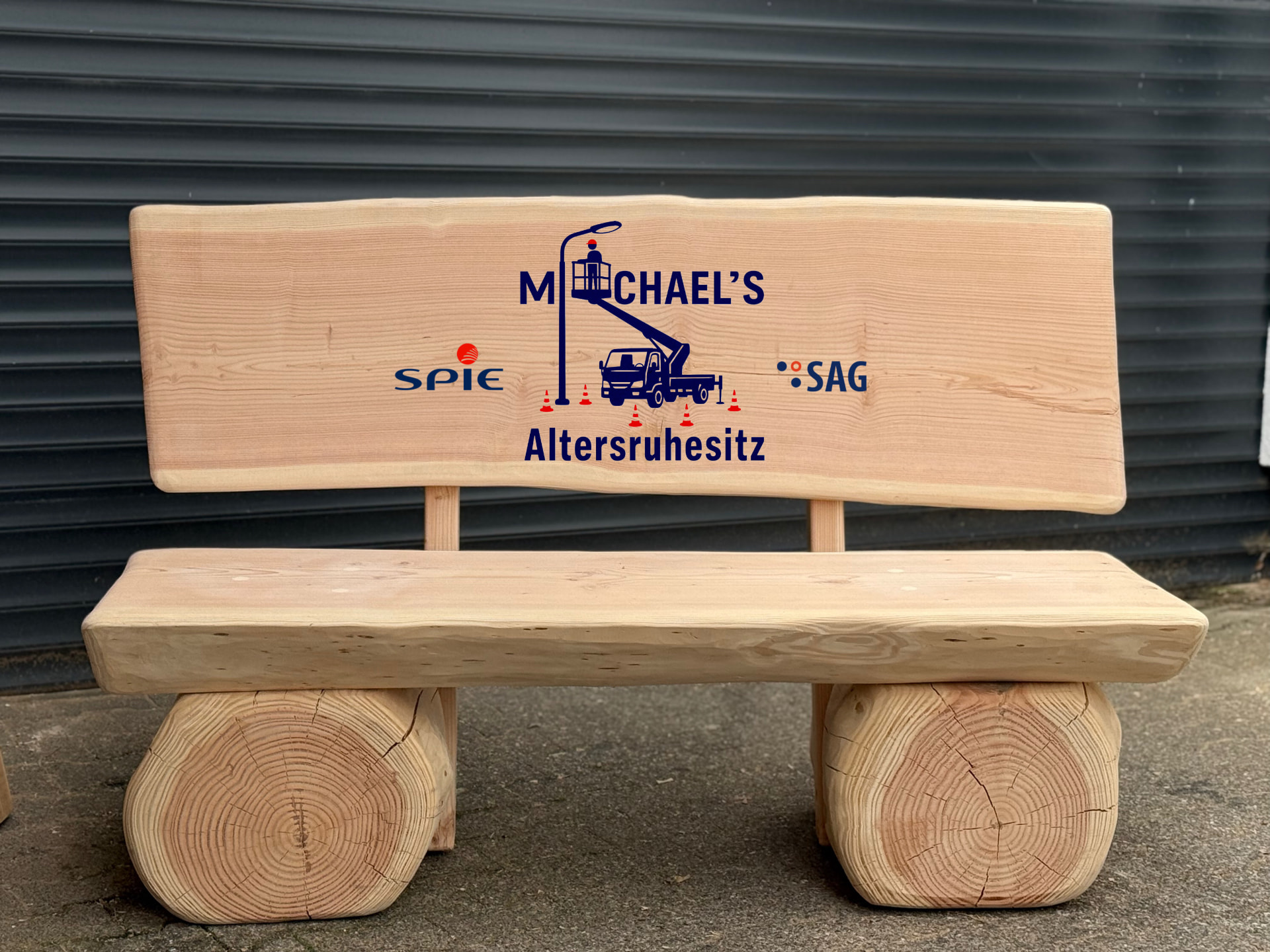 Individuell gefertigte massive Holzbank mit farbig ausgearbeiteter CNC-Gravur „Michael's Altersruhesitz" sowie den Firmenlogos von SPIE und SAG, personalisiert als besonderes Geschenk für einen Mitarbeiter oder Kollegen.