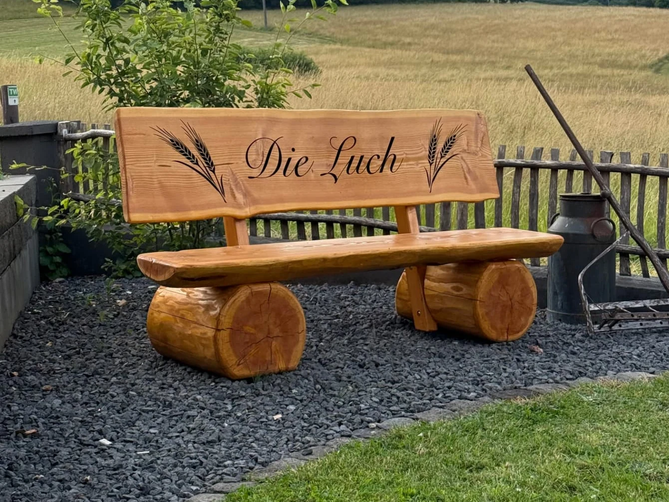 Baumstammbank mit CNC-Gravur 'Die Luch' und Ährenmotiven Baumstammbank mit schwarz ausgearbeiteter CNC-Gravur 'Die Luch' und Ährenmotiven vor einem Feld.