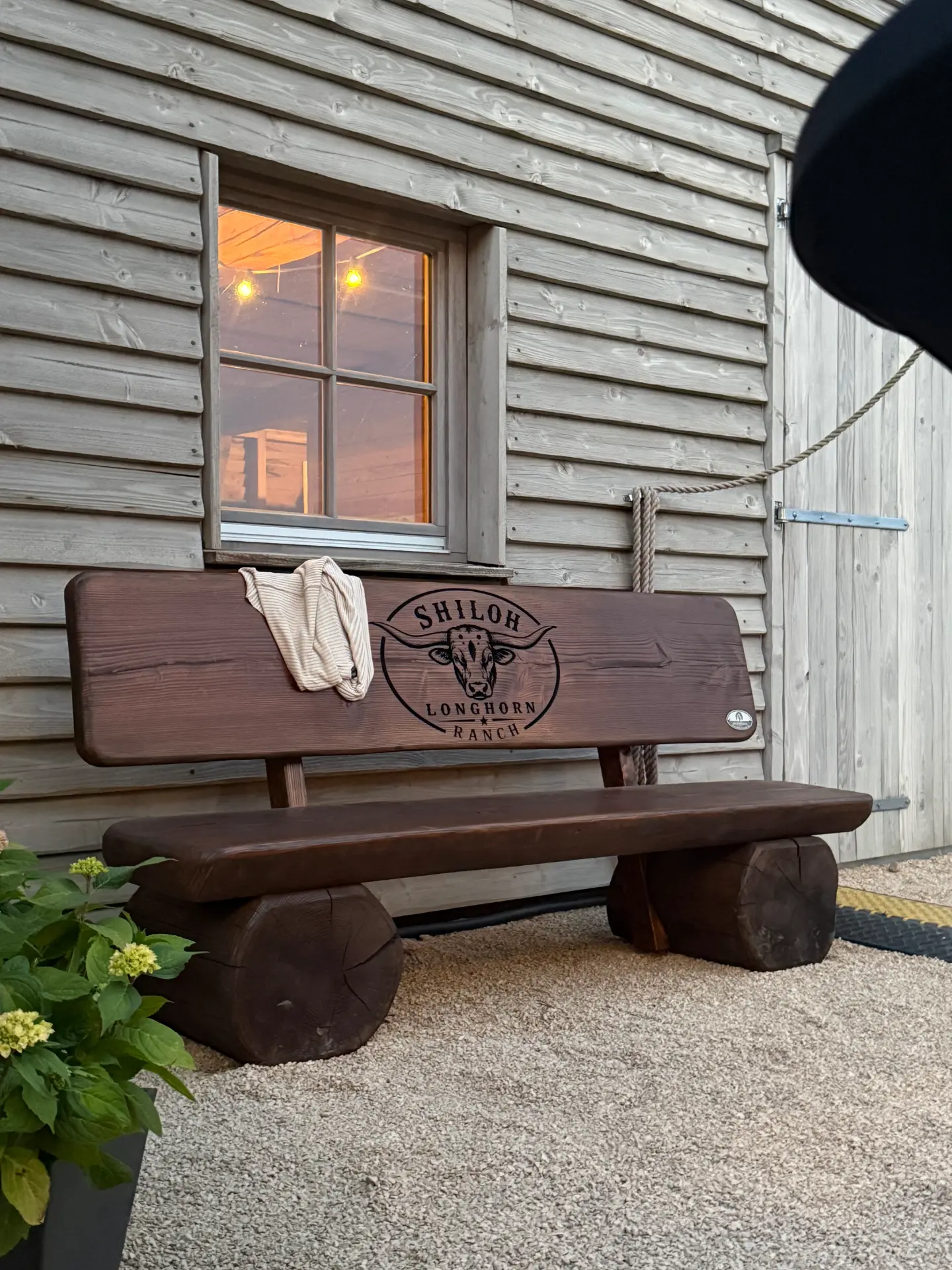 Personalisierte Baumstammbank mit CNC-gefrästem Longhorn-Ranch-Logo Personalisierte Baumstammbank in 220cm Sitzbreite, mit CNC-gefrästem Shiloh-Longhorn-Ranch-Logo vor einem Holzgebäude